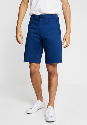 Homme portant un short bleu mi-genoux, des baskets blanches et un t-shirt blanc, debout devant un fond clair uni.