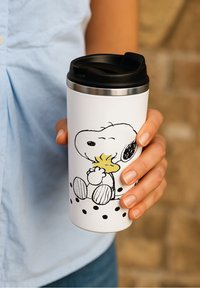 Una mano tiene una tazza da viaggio bianca con coperchio nero che presenta Snoopy che abbraccia il cartone giallo Woodstock, su uno sfondo sfocato beige.