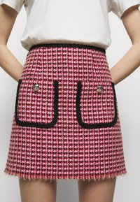 Mini-jupe en tweed rouge et rose avec bordure noire, comprenant deux poches avant avec boutons décoratifs et un ourlet effiloché.
