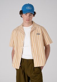 Short-sleeve shirt with yellow-orange vertical stripes, beige base, and embroidered logo; paired with a blue cap and olive green trousers.
