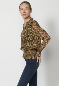Blusa nera a fiori con fiori gialli e arancioni, maniche lunghe e vita arricciata. Abbinata a jeans blu scuro.