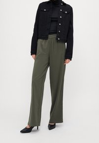 Black denim jacket with silver buttons, paired with olive green elastic-waist wide-leg pants and black pointed-toe heels.