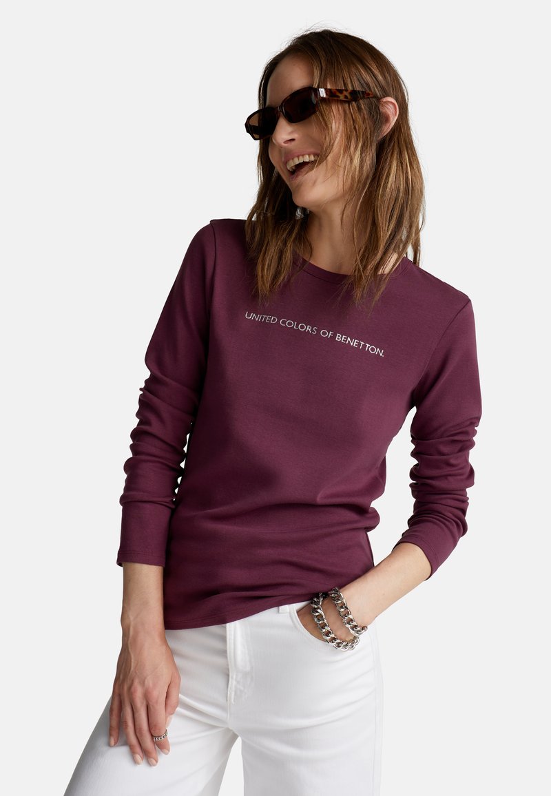 Burgundowa koszula z długim rękawem z napisem "UNITED COLORS OF BENETTON", w połączeniu z białymi spodniami i biżuterią srebrną. Teksturowana tkanina.