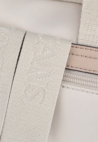 Borsa in tessuto beige con una superficie liscia, dotata di zip e ampia tracolla con dettagli in rilievo del logo.