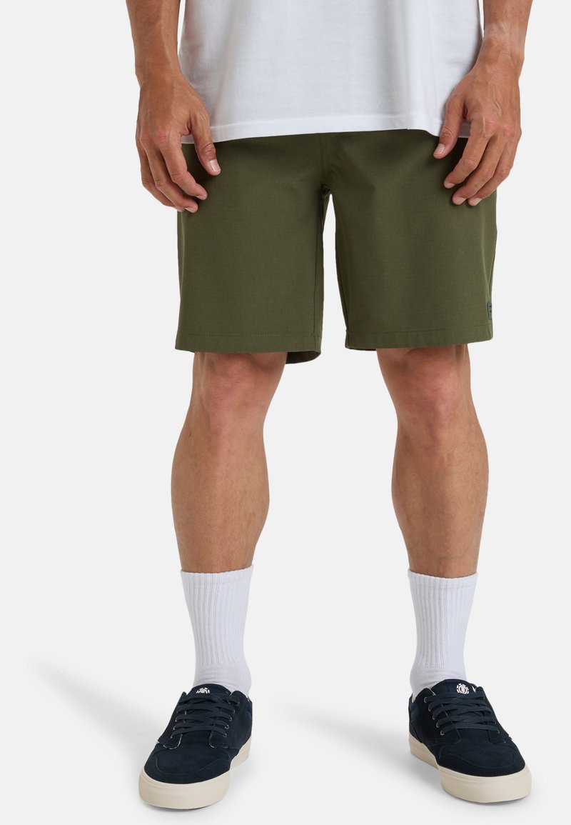 Pantaloni corti verde oliva con parte frontale piatta, tasche laterali e dettaglio del logo. Indossati con calzini bianchi a costine e scarpe di camoscio blu navy.