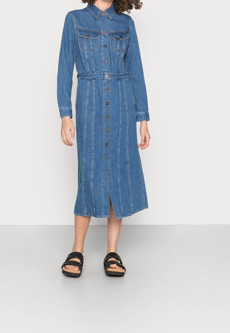 Robe chemise en denim à manches longues, col, fermeture à boutons sur le devant et deux poches poitrine. Présente des rayures verticales et une fente jusqu'au genou. Portée avec des sandales noires.