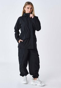 Giacca leggera nera con colletto alto e cordoncino, dotata di vita cinzata e tasche laterali, abbinata a pantaloni cargo neri.