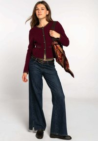 Cárdigan burdeos con ribetes verdes y bolsillos delanteros, combinado con jeans de mezclilla oscuros de pierna ancha. La modelo sostiene una bufanda estampada y un bolso.