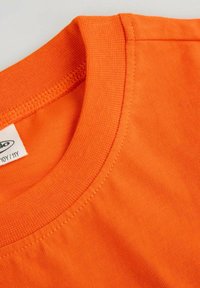 Zbliżenie na pomarańczową koszulkę z okrągłym dekoltem, ukazującą fakturę materiału, szwy oraz częściowo widoczną białą metkę odzieżową.