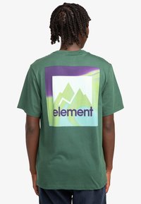 Camiseta de algodón verde con un gran gráfico de montañas en tonos azules y verdes, con la palabra "elemento" en púrpura en negrita en la parte central de la espalda.