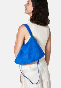 Blå mocka axelväska med en triangulär form; har en guldfärgad logotyp och en texturerad yta. Lång blå rem med guldfärgad hårdvara.