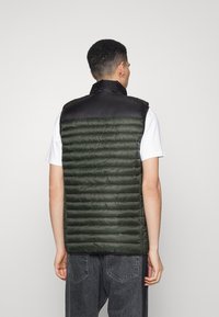 Chaleco acolchado con una sección superior negra y una sección inferior de color verde oscuro acolchada. Cuenta con cierre de cremallera y sin mangas, superficie texturizada con líneas horizontales.