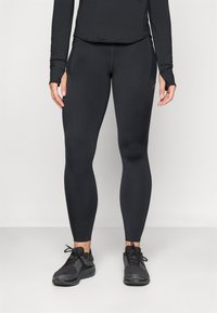 Kvinne iført svarte tettsittende trenings tights og svarte joggesko, stående mot en enkel lys bakgrunn.