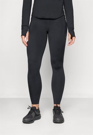 Kvinne iført svarte tettsittende trenings tights og svarte joggesko, stående mot en enkel lys bakgrunn.