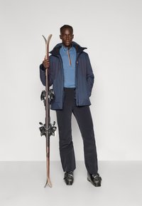 Skiausrüstungsdisplay: eine Person in einer blauen und marineblauen Jacke, blauer Unterwäsche, schwarzen Skihosen, die Holzskier mit schwarzen Bindungen hält.