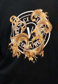 Λογότυπο Versace Jeans Couture με λευκά γράμματα, περικυκλωμένο από περίτεχνες χρυσές στροβιλισμένες διακοσμήσεις μπαρόκ σε μαύρο υφασμάτινο φόντο.