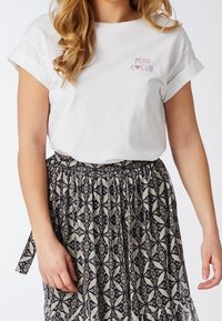 Mujer vistiendo una camiseta blanca con el texto "Plan Coeur" y una falda negra y beige estampada con un diseño floral geométrico.