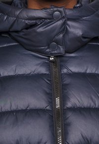 Marinblå pufferjacka med hög krage, tryckknappsstängningar och en central dragkedja. Materialet verkar slätt med quiltade sektioner.