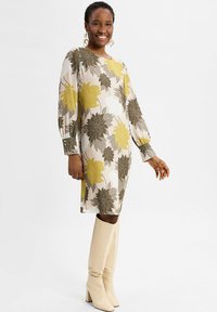 Lächelnde Frau in einem langärmeligen Blumenkleid mit gelben, braunen und weißen Blumen, kombiniert mit kniehohen cremefarbenen Stiefeln.