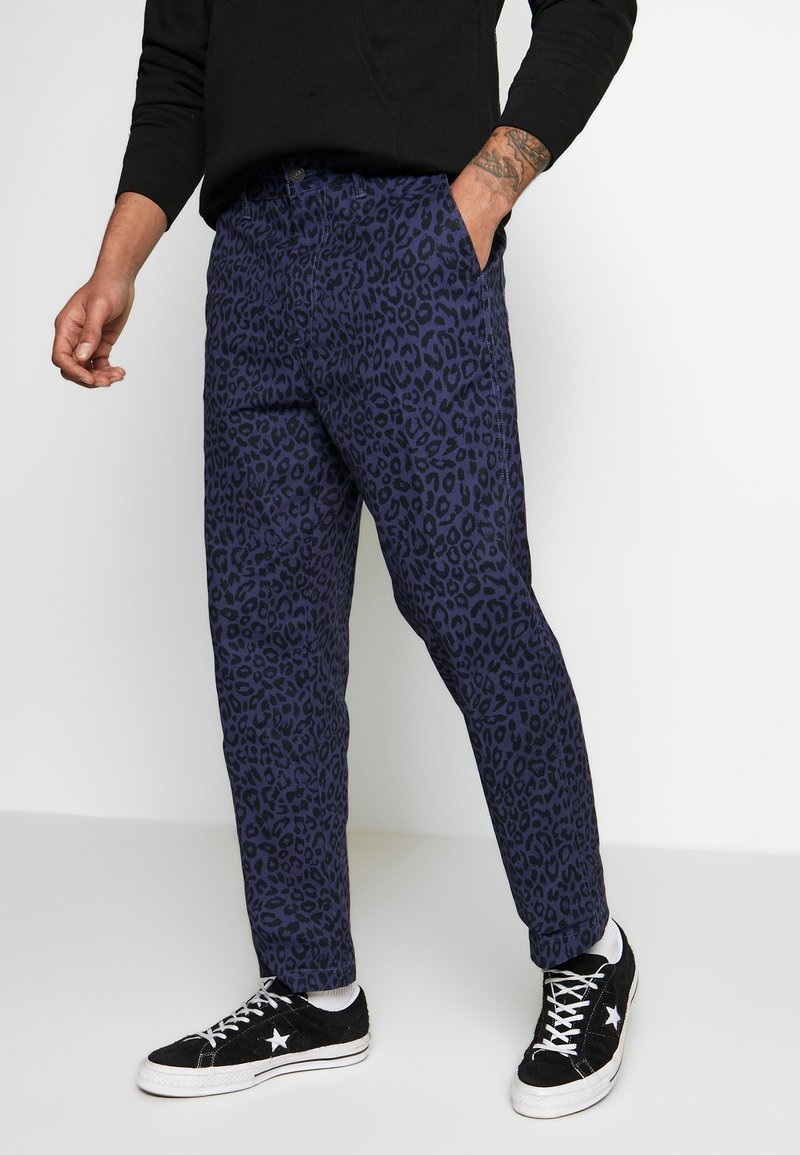 Muž v tmavě modrých kalhotách s černým leopardím potiskem, v černých teniskách s bílou hvězdou a bílých ponožkách, stojící na bílé podlaze s jednou rukou v kapse.