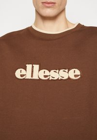 Sudadera marrón con un logo "ellesse" bordado en beige. Cuello redondo con un ribete acanalado beige en contraste. Textura de algodón suave.
