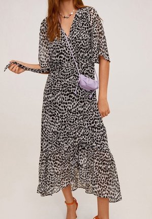 Kobieta ubrana w czarno-białą sukienkę midi w panterkę z dekoltem w serek, trzyma pasek, ma małą fioletową torebkę na ramię i pomarańczowe sandały na obcasie.