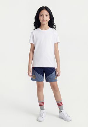 Barn med langt mørkt hår står og vender fremad iført en enkel hvid T-shirt, blå sportsshorts, grå sokker med røde striber og hvide sneakers.