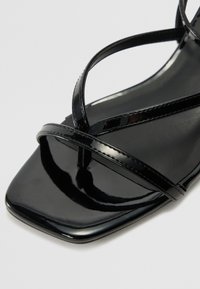 Sandale en cuir verni noir avec un orteil carré, dotée de plusieurs fines lanières entrecroisées soutenant le pied et d'une surface lisse et brillante.