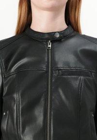 Giacca di pelle nera con colletto a camicia, zip frontale e due tasche sul petto. Texture liscia con dettagli cuciti.