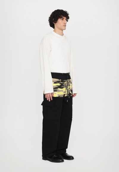 Top alb cu mâneci lungi, pantaloni de tip cargo negri cu accente de camuflaj galben, având buzunare și talie cu șnur. Pantofi negri completează ținuta.