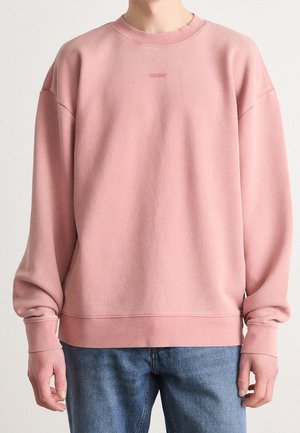 Osoba ubrana w luźną, pudrowo-różową bluzę z subtelnym logo "HUGO" na piersi oraz niebieskie dżinsy, stojąca na jednolitym tle.