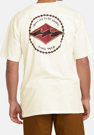 Personne portant un t-shirt crème avec un logo rond sur le thème du surf dans le dos indiquant "Quality Surf Goods Since 1973" et un short marron.