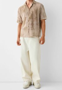 Beige shirt met korte mouwen en een netwerkpatroon, gecombineerd met losse witte broek en lichtgekleurde sneakers.