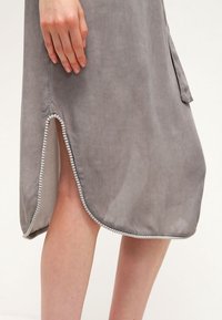 Harmaasta mokkanahasta valmistettu mekko, jossa on kaareva helma, sivuventura ja kontrastiväri valkoista helminauhaa reunan ympärillä. Sileä pinta.