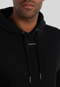 Felpa con cappuccio nera realizzata in tessuto morbido, con logo frontale, cordini regolabili e dettagli metallici argentati alle punte.