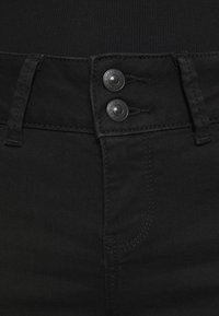 Černé džíny z denimu s obepnutým pasem, které mají dva černé knoflíky, jemné švy a hladkou texturu. Viditelný detail zapínání na sponu.