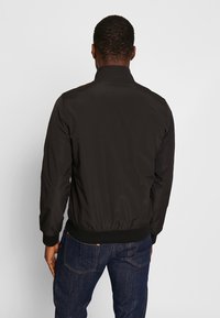 Sort bomberjakke med ståkrave, elastiske manchetter og kant, har en glat tekstur og minimalistisk design. Båret med mørke jeans.