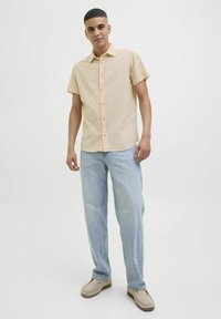 Lichtbeige overhemd met korte mouwen en knopen, gecombineerd met lichtblauwe recht gesneden jeans en beige instappers. Gladde stoftextuur.