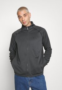 Giacca sportiva nera zip-up con colletto alto, caratterizzata da una texture liscia e dal logo Nike. Il modello indossa jeans blu.