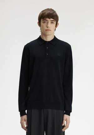 Giovane uomo con capelli castani corti che indossa una polo nera a maniche lunghe e pantaloni scuri, in piedi davanti a uno sfondo chiaro e uniforme.
