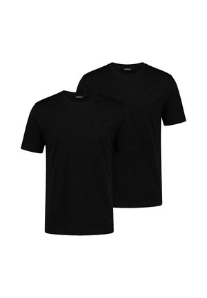 Hombre con el pelo corto y barba clara que lleva una camiseta negra lisa de manga corta y pantalones blancos, de pie frente a un fondo gris.