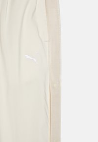 Pantalones deportivos beige de tejido suave, con un logo de Puma bordado en blanco y detalles de botones a presión a lo largo de las costuras laterales.