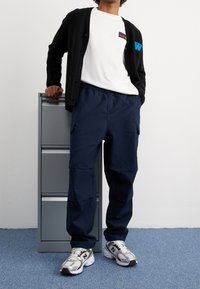 Czarny kardigan, biały T-shirt z logo, granatowe spodnie cargo, szare sneakersy. Gładkie tkaniny i luźny krój, z funkcjonalnymi bocznymi kieszeniami.