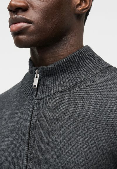 Sivý pletený sveter s vysokým rebrovaným golierom a strieborným zipsom. Má textúrovaný povrch a jemné detaily švov.