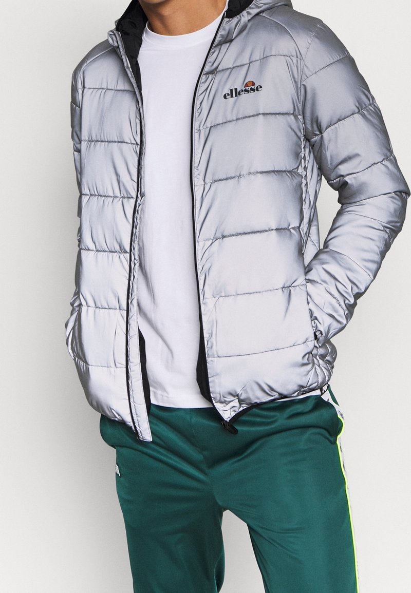 Homme portant une veste matelassée argentée avec le logo ellesse, un t-shirt blanc et un pantalon de survêtement vert avec une bande latérale jaune, les mains dans les poches de la veste.