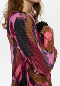 Krāsains plisēts blūze ar drosmīgiem svītrām rozā, brūnā un melnā krāsā. Garas piedurknes ar ribotiem melniem aprocēm. Tekstūrēta auduma dizains.