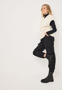 Gilet en peluche blanc sur une chemise à manches longues noire, associé à un pantalon cargo noir et des bottines noires. Textures lisses et design pratique.