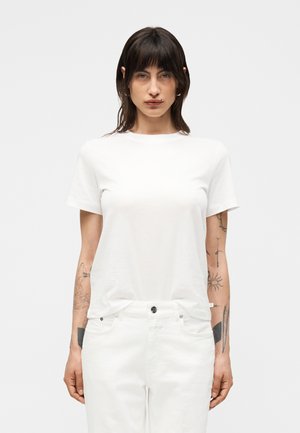 Kobieta o ciemnych włosach, ubrana w prosty biały t-shirt z krótkim rękawem i białe spodnie, stoi z widocznymi tatuażami na obu przedramionach.