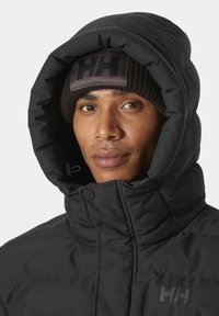 Chaqueta acolchada negra con cuello alto y capucha ajustable. Presenta un parche con logo en un gorro de punto oscuro. Textura suave y mate.