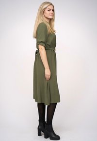 Rochie midi de culoare verde măslinie cu mâneci scurte pufoase și talie împregnată. Purtați cu botine negre. Material neted și siluetă lejeră.